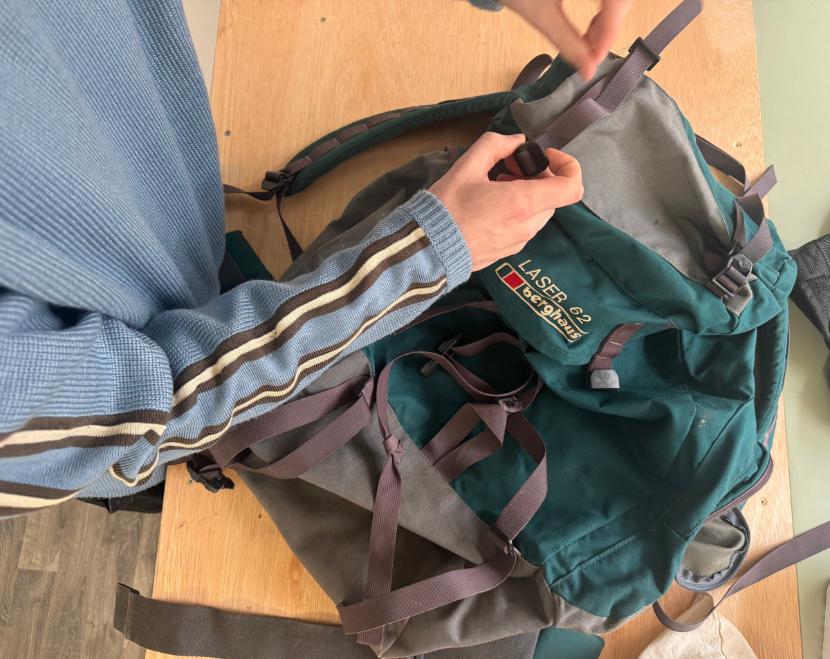 A person wearing a blue striped sweater is adjusting the straps of a green and gray backpack on a wooden table. The backpack has a visible 'LASER CRAFT' logo patch.