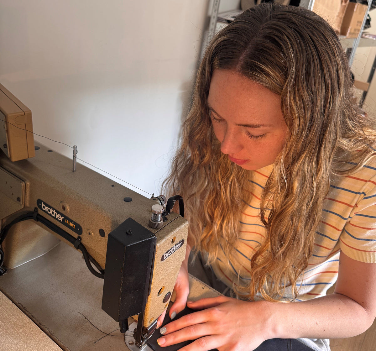 A woman with long wavy blonde hair operates a Brother industrial sewing machine in a workshop setting.