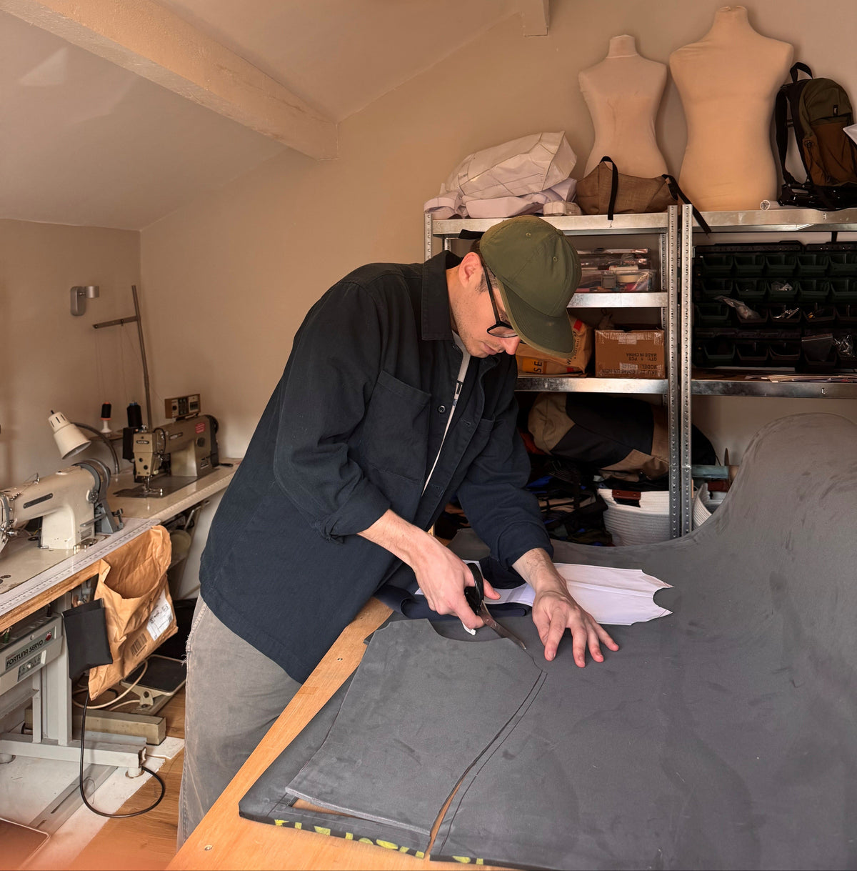 A person in a dark jacket and green cap is cutting a large gray fabric piece on a table in a sewing room with shelves, mannequins, and sewing machines visible.