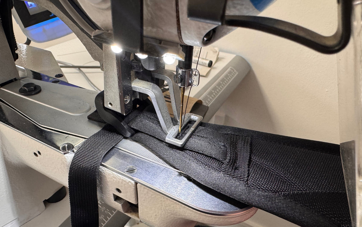 A close-up of an industrial sewing machine stitching a black fabric strap or trim, with the needle, presser foot, and machine body clearly visible.